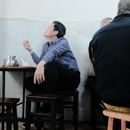 A person in a blue checkered shirt is seated on a wooden stool, holding a cigarette in one hand. The setting appears to be indoors, with plain white tile walls. A metal teapot and a small cup are on the table.