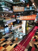 A modern shopping mall interior with a vibrant and colorful design. Large signs with 'I ❤️ ART' and '#FUNANSG' are suspended above an open area. The floor features an eye-catching geometric pattern in various shades. A red path marked for cycling and walking crosses through, displaying instructions to slow down. Escalators connect the different levels, and the space is filled with a mix of natural and artificial lighting.