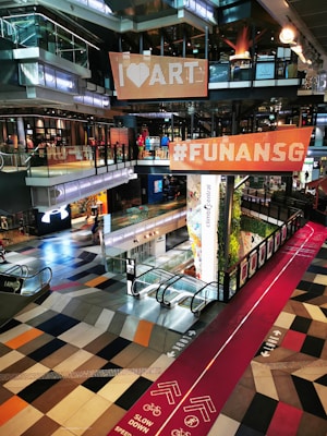 A modern shopping mall interior with a vibrant and colorful design. Large signs with 'I ❤️ ART' and '#FUNANSG' are suspended above an open area. The floor features an eye-catching geometric pattern in various shades. A red path marked for cycling and walking crosses through, displaying instructions to slow down. Escalators connect the different levels, and the space is filled with a mix of natural and artificial lighting.