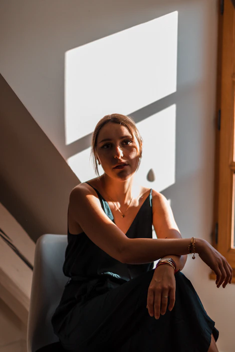 A stylish woman wearing minimalist, elegant clothing in neutral tones, standing in a softly lit modern room with gold accents.