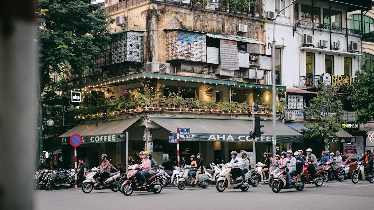Carrefour animé d'une ville d'Asie du Sud-Est, avec des dizaines de scooters devant des façades anciennes et un café