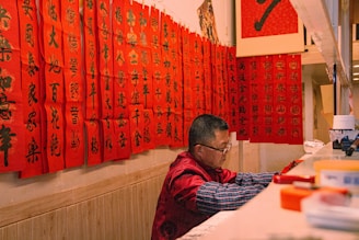 A man is seated at a counter, deeply focused on his work. The wall is adorned with numerous red banners featuring Chinese calligraphy in black ink. The workspace is filled with various objects including containers and writing tools. The setting conveys a traditional and cultural ambiance.