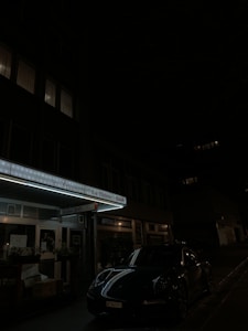 A dark street view featuring a building with illuminated signs, including 'Le Velours' and 'Le Bidou BAR'. A sports car is parked on the dimly lit street, and some windows in the building are faintly glowing from interior lights.