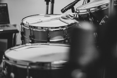 A close-up, black and white photograph of a drum kit featuring a snare drum with drumsticks resting on top. A cloth lies draped across the snare, and nearby are parts of additional drums and stands, with microphones positioned overhead for recording. In the background, part of a laptop is visible.