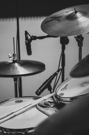 A black and white photograph of a drum set featuring cymbals and a snare drum. Drumsticks rest on the snare drum, and a microphone is positioned nearby, suggesting a studio or recording setting. The overall setup indicates a focus on percussion instruments.