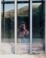 A reflection of a person holding a camera is visible through a dusty window with metal bars. The background appears to be an outdoor landscape with hills. The image captures the silhouette of the photographer amidst natural and rustic elements.