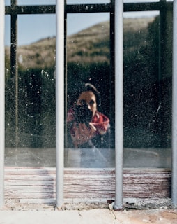 A reflection of a person holding a camera is visible through a dusty window with metal bars. The background appears to be an outdoor landscape with hills. The image captures the silhouette of the photographer amidst natural and rustic elements.