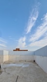 An open rooftop area enclosed by a corrugated metal fence, with a tiled floor and some scattered construction materials and debris. In the distance, a red brick building is visible against a clear blue sky with soft, wispy clouds.