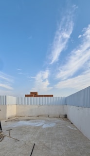 An open rooftop area enclosed by a corrugated metal fence, with a tiled floor and some scattered construction materials and debris. In the distance, a red brick building is visible against a clear blue sky with soft, wispy clouds.