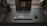A sleek, modern setup featuring a laptop with the screen displaying a logo for MAR Gaming. The laptop is placed on a clean, dark desk, accompanied by a gaming mouse on the right and a pair of headphones resting on a stand to the left. A microphone is positioned on the right side of the desk.