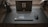 A sleek, modern setup featuring a laptop with the screen displaying a logo for MAR Gaming. The laptop is placed on a clean, dark desk, accompanied by a gaming mouse on the right and a pair of headphones resting on a stand to the left. A microphone is positioned on the right side of the desk.
