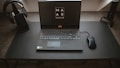 A sleek, modern setup featuring a laptop with the screen displaying a logo for MAR Gaming. The laptop is placed on a clean, dark desk, accompanied by a gaming mouse on the right and a pair of headphones resting on a stand to the left. A microphone is positioned on the right side of the desk.