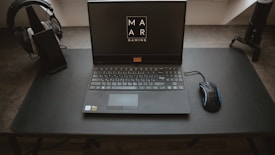 A sleek, modern setup featuring a laptop with the screen displaying a logo for MAR Gaming. The laptop is placed on a clean, dark desk, accompanied by a gaming mouse on the right and a pair of headphones resting on a stand to the left. A microphone is positioned on the right side of the desk.