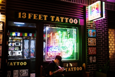 A tattoo shop with a prominent neon sign reading '13 Feet Tattoo' in glowing colors. The storefront features a large window displaying colorful tattoo designs and there is art also displayed on the exterior wall. A person is seated outside the shop, possibly browsing on a phone.