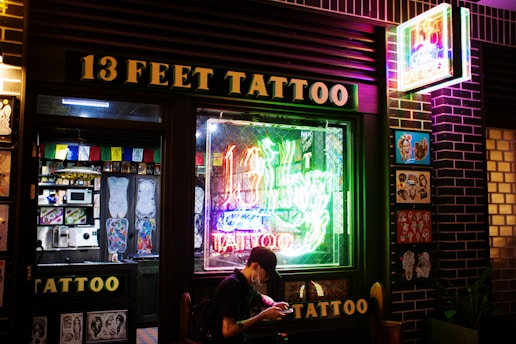 A tattoo shop with a prominent neon sign reading '13 Feet Tattoo' in glowing colors. The storefront features a large window displaying colorful tattoo designs and there is art also displayed on the exterior wall. A person is seated outside the shop, possibly browsing on a phone.