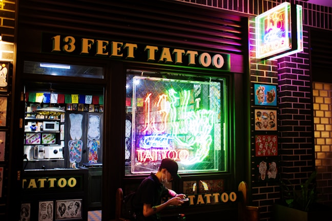 A tattoo shop with a prominent neon sign reading '13 Feet Tattoo' in glowing colors. The storefront features a large window displaying colorful tattoo designs and there is art also displayed on the exterior wall. A person is seated outside the shop, possibly browsing on a phone.