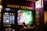 A tattoo shop with a prominent neon sign reading '13 Feet Tattoo' in glowing colors. The storefront features a large window displaying colorful tattoo designs and there is art also displayed on the exterior wall. A person is seated outside the shop, possibly browsing on a phone.