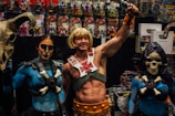 A group of people dressed in elaborate costumes inspired by fantasy characters, including a muscular figure holding a sword and two companions wearing makeup and thematic outfits. They are in front of a display filled with action figures and related merchandise.