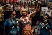 A group of people dressed in elaborate costumes inspired by fantasy characters, including a muscular figure holding a sword and two companions wearing makeup and thematic outfits. They are in front of a display filled with action figures and related merchandise.