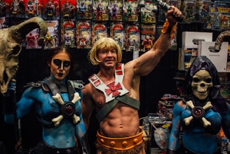 A group of people dressed in elaborate costumes inspired by fantasy characters, including a muscular figure holding a sword and two companions wearing makeup and thematic outfits. They are in front of a display filled with action figures and related merchandise.