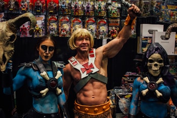 A group of people dressed in elaborate costumes inspired by fantasy characters, including a muscular figure holding a sword and two companions wearing makeup and thematic outfits. They are in front of a display filled with action figures and related merchandise.