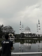 A person wearing a shirt with the word 'TAIM' stands near a fence overlooking a reflective body of water. Across the water, a large mosque with four tall minarets and a distinctive blue and white dome is surrounded by trees under a cloudy sky.