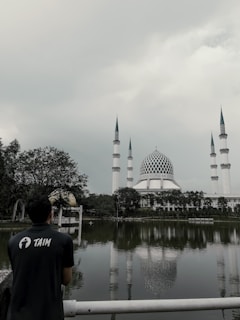 A person wearing a shirt with the word 'TAIM' stands near a fence overlooking a reflective body of water. Across the water, a large mosque with four tall minarets and a distinctive blue and white dome is surrounded by trees under a cloudy sky.
