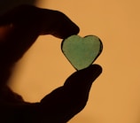 A silhouette of a hand holding a translucent, heart-shaped object against a warm, golden background. The heart appears to be made of glass or resin with small air bubbles or imperfections visible inside.