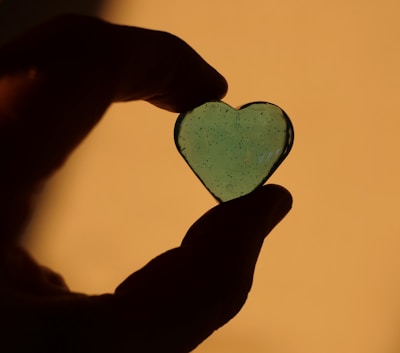 A silhouette of a hand holding a translucent, heart-shaped object against a warm, golden background. The heart appears to be made of glass or resin with small air bubbles or imperfections visible inside.