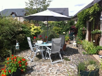 A serene garden patio setting with a circular glass table and four gray chairs under a large gray umbrella. The area is surrounded by lush greenery and vibrant flowers in pots. The stone pathway adds a rustic touch, and there are decorative lanterns placed around the scene. A quaint house with stone walls and a slate roof is visible in the background.