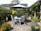 A serene garden patio setting with a circular glass table and four gray chairs under a large gray umbrella. The area is surrounded by lush greenery and vibrant flowers in pots. The stone pathway adds a rustic touch, and there are decorative lanterns placed around the scene. A quaint house with stone walls and a slate roof is visible in the background.