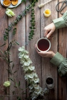 A rustic wooden table with a person's hands holding a cup of tea. Nearby are white flowers and green foliage arranged decoratively. A small honey jar with a honey dipper, some cookies on a plate, and a pair of scissors are visible. The scene is softly lit, creating a cozy and serene atmosphere.