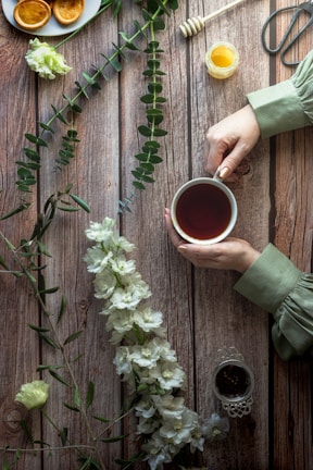 A rustic wooden table with a person's hands holding a cup of tea. Nearby are white flowers and green foliage arranged decoratively. A small honey jar with a honey dipper, some cookies on a plate, and a pair of scissors are visible. The scene is softly lit, creating a cozy and serene atmosphere.