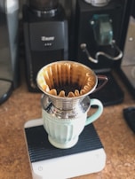 A coffee brewing setup featuring a metal pour-over dripper placed atop a light green ceramic mug. The setup rests on a digital scale, surrounded by coffee-making equipment like grinders and coffee machines.