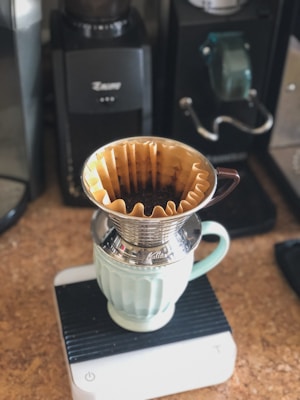 A coffee brewing setup featuring a metal pour-over dripper placed atop a light green ceramic mug. The setup rests on a digital scale, surrounded by coffee-making equipment like grinders and coffee machines.