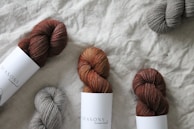 Several skeins of yarn are arranged on a textured fabric surface. The yarn colors include shades of brown and gray, and some are wrapped with labels that read 'Seasons.' The yarn has a soft, cozy texture.