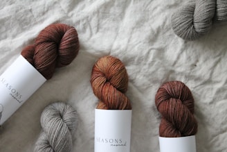 Several skeins of yarn are arranged on a textured fabric surface. The yarn colors include shades of brown and gray, and some are wrapped with labels that read 'Seasons.' The yarn has a soft, cozy texture.