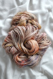 A large bundle of multicolored yarn is artfully arranged on a soft, light-colored fabric. The yarn features a mix of beige, pink, orange, and hints of blue, creating a visually appealing texture.