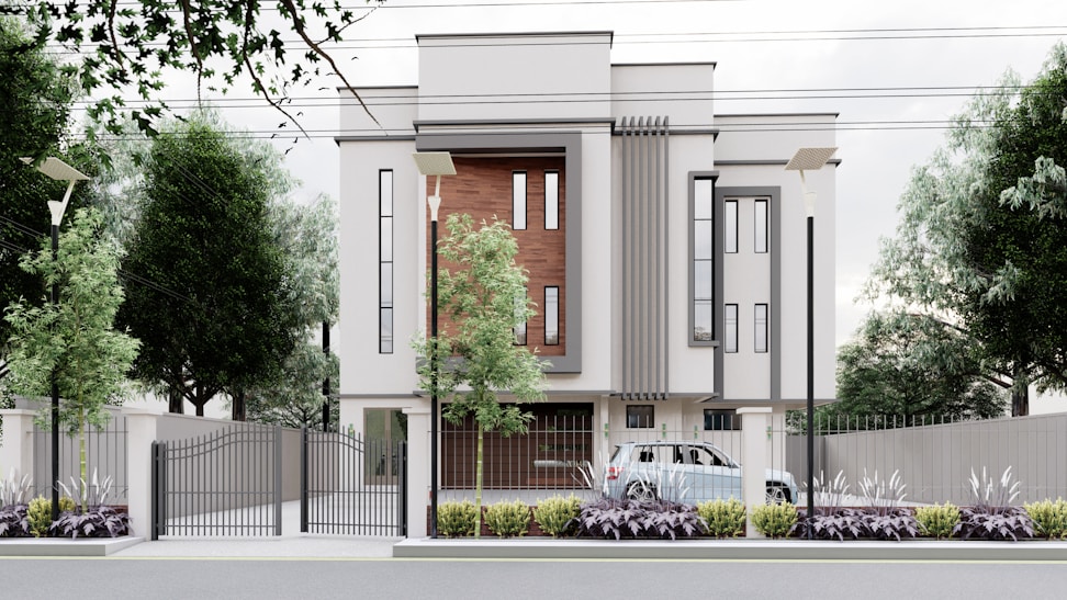 A modern, multi-story building with a minimalist design features large vertical windows and accent walls with wooden panels. The property is surrounded by a metal fence with a gate, and there are street lamps and tall trees along the perimeter. A car is parked inside the gated area, and the landscaped front garden is adorned with shrubs and plants.