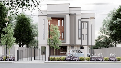 A modern, multi-story building with a minimalist design features large vertical windows and accent walls with wooden panels. The property is surrounded by a metal fence with a gate, and there are street lamps and tall trees along the perimeter. A car is parked inside the gated area, and the landscaped front garden is adorned with shrubs and plants.