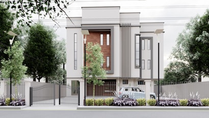 A modern, multi-story building with a minimalist design features large vertical windows and accent walls with wooden panels. The property is surrounded by a metal fence with a gate, and there are street lamps and tall trees along the perimeter. A car is parked inside the gated area, and the landscaped front garden is adorned with shrubs and plants.