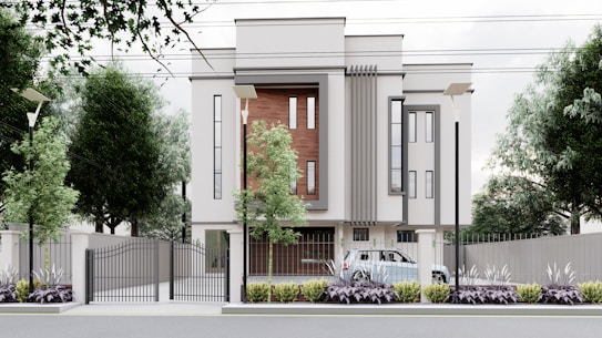 A modern, multi-story building with a minimalist design features large vertical windows and accent walls with wooden panels. The property is surrounded by a metal fence with a gate, and there are street lamps and tall trees along the perimeter. A car is parked inside the gated area, and the landscaped front garden is adorned with shrubs and plants.