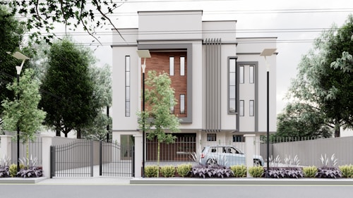 A modern, multi-story building with a minimalist design features large vertical windows and accent walls with wooden panels. The property is surrounded by a metal fence with a gate, and there are street lamps and tall trees along the perimeter. A car is parked inside the gated area, and the landscaped front garden is adorned with shrubs and plants.