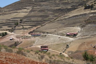 A rural landscape with terraced hills. Several small houses with red roofs are scattered along dirt roads in a dry, barren terrain. Sparse vegetation and winding paths are evident.