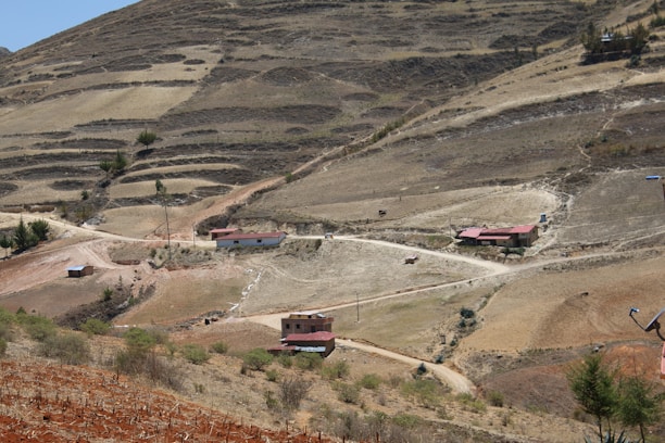 A rural landscape with terraced hills. Several small houses with red roofs are scattered along dirt roads in a dry, barren terrain. Sparse vegetation and winding paths are evident.