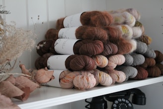 A neatly arranged stack of yarn skeins in various earthy and muted tones, including browns, grays, and creams, placed on a white shelf. Dried leaves and flowers add a rustic touch to the composition, while an old-fashioned rotary phone sits beneath the shelf, adding a vintage element.