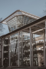 A modern architectural structure features expansive glass panels and angular lines, creating a sleek and reflective facade. The design incorporates a large, tapered glass tower at the center, surrounded by metal frameworks and supports.