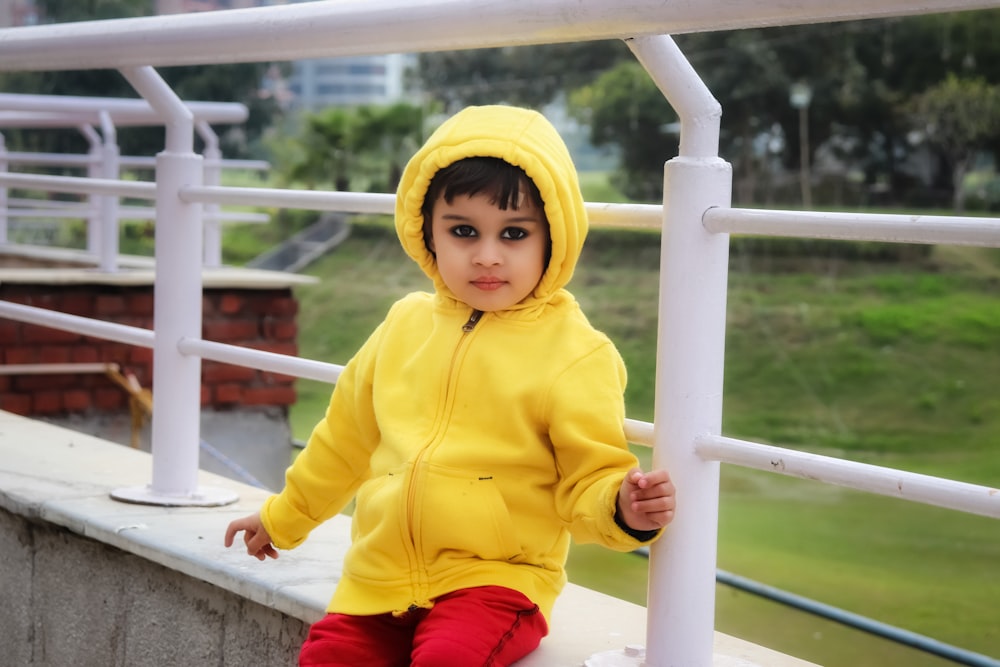child in yellow hoodie jacket and red pants sitting on white metal railings during daytime