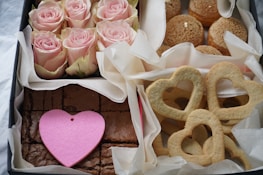 An assortment of items arranged neatly in a box lined with white paper. The top left section contains several pink roses. To the right, there are round pastries with a crumbly top. In the lower right are heart-shaped cookies. The bottom left has chocolate brownies topped with a large pink heart decoration.