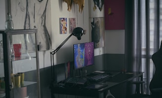 A dimly lit room features a desk with a computer displaying colorful artwork on the screen. On the wall are various art pieces, including abstract and figure drawings. A black desk lamp arches over the computer. To the left, a glass cabinet holds multiple mugs, one of which is red with white text. Curtains filter light in the background.